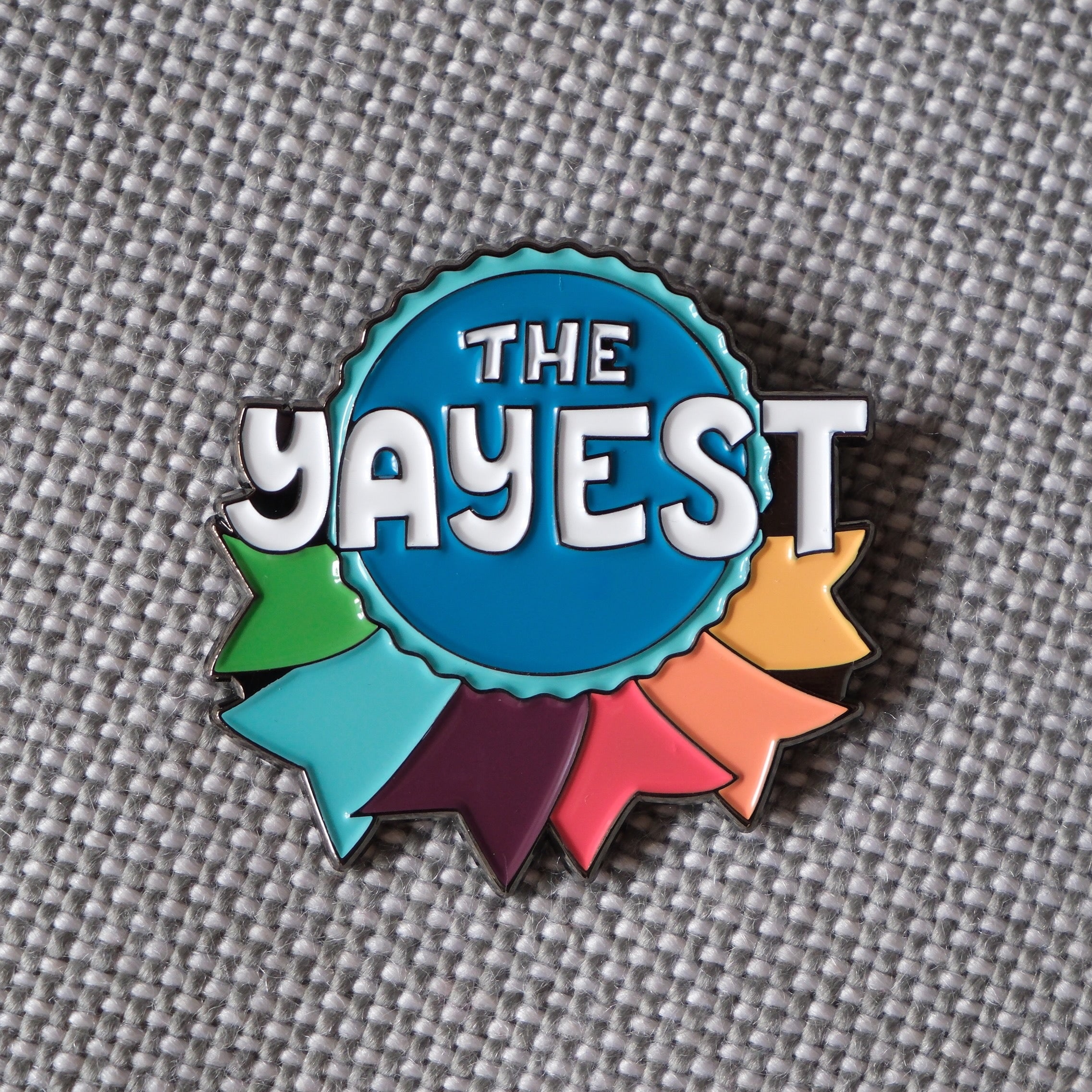 Detail of an enamel pin shaped like a ribbon and reads The Yayest