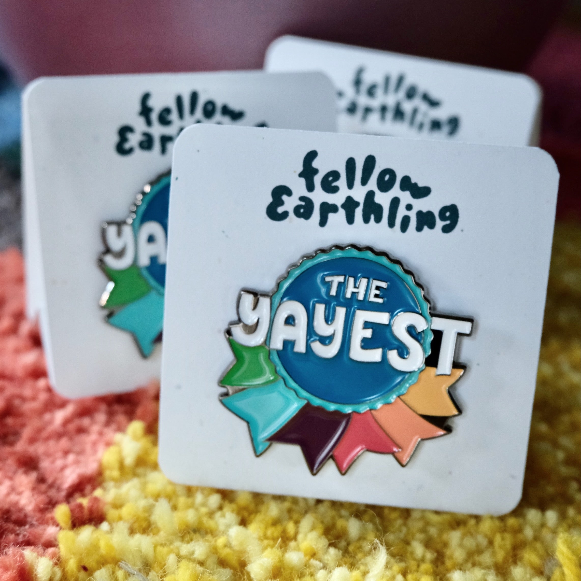Detail of an enamel pin shaped like a ribbon and reads The Yayest